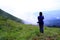 Asian hiking or tourist woman in dark blue sweater with camera standing on green grass field with mountain view and mist