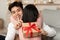 Asian Husband Holding Gift Gesturing Hush Embracing Girlfriend Sitting Indoors