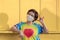 Asian LGBT woman in rainbow t-shirt with protective mask looking at camera with showing love hand sign on wooden yellow background
