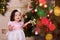 Asian little cute girl wearing casual dress posing close to new year christmas green classic tree with balls toys and colourful pr