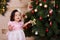 Asian little cute girl wearing casual dress posing close to new year christmas green classic tree with balls toys and colourful pr