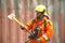 An Asian male fireman in orange protective clothing, mask and helmet with an ax standing front of building.