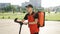 Asian man courier food delivery with red thermal backpack walks street with electric scooter uses smartphone navigate