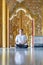 Asian Man meditates on the ground with reflection in the sunlight pouring into the temple church with gold gold Thai pattern blur