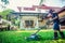 Asian man mowing lawn in the garden with blurry house. Mowing or cutting the long grass with electric lawn mower