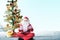 Asian man in Santa costume with open palm gesture sitting with Christmas tree