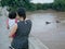 Asian mother carrying her little baby girl standing on a bridge watching a long-tail boat passing by on the river