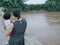 Asian mother carrying her little daughter standing on a bridge showing her little baby a murky muddy river aft