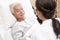 Asian old man lying in bed talking to young female doctor in hospital ward