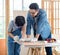 Asian professional male carpenter woodworker engineer dad in jeans outfit with safety gloves goggles helping teaching young boy