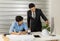 Asian senior good-looking boss in suit sitting at desk and working with junior employee in office with roll of paper. Businessmen