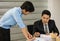 Asian senior good-looking boss in suit sitting at desk and working with junior employee in office with roll of paper. Businessmen