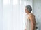 Asian senior woman white hair standing with walking crutches near curtain looking out glass window.