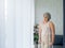 Asian senior woman white hair standing with walking crutches near curtain looking out glass window.