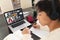 Asian teenage boy writing notes in book while attending online class over video call on laptop