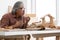 Asian white haired senior carpenter man blowing sawdust and using sandpaper to smooth wooden model at home workshop