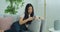 Asian woman chatting use smartphone sitting on comfortable couch in living-room. Young female smiling surfing internet