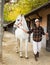 Asian woman leading white horse by bridle outdoors along stables