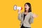Asian woman screaming in a megaphone spreading her hands looking away on a bright yellow background