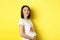 Asian woman smiling kindly, touching belly and looking at camera, being pregnant, having a baby, standing over yellow