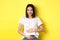 Asian woman smiling kindly, touching belly and looking at camera, being pregnant, having a baby, standing over yellow