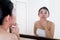 Asian woman squeezing pimples on her face while looking at reflection in mirror.