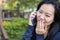 Asian woman talking on smart phone and holding her hand against her mouth at park. Communication concept. Extrovert characteristic