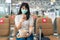 Asian woman tourist wearing mask using mobile phone searching airline flight status and sitting social distancing chair in airport