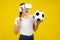 Asian woman in VR glasses and holding a soccer ball, isolated yellow wall background, Sport and Metaverse