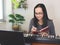 Asian woman wearing eyeglasses sitting with electric piano, writing on notebook looking at computer notebook on table. Teaching or