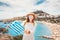 Asian woman in white dress posing with greek flag in front of the famous Lindos town on Rhodes island. Vacation and travel