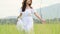 Asian woman in white dress is strolling in vast grassland and touching top of green grass by hand. Girl walking in meadow with