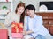 Asian young beautiful female girlfriend sitting smiling opening unboxing red present gift wrapped box on cozy sofa from handsome