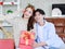 Asian young beautiful female girlfriend sitting smiling opening unboxing red present gift wrapped box on cozy sofa from handsome