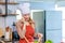 Asian young female chef housewife wears white tall cook hat and apron standing smiling on smartphone call holding wooden spoon