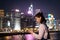 Asian young lady with face mask messages and reads on mobile phone with blurred Colorful magnificent Night city view of Central,