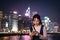 Asian young lady with face mask messages and reads on mobile phone with blurred Colorful magnificent Night city view of Central,