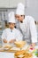 Asian young little boy pastry chef in white uniform with tall cook hat standing while male cooking teacher smiling help teaching