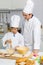 Asian young little boy pastry chef in white uniform with tall cook hat standing while male cooking teacher smiling help teaching