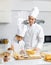 Asian young little boy pastry chef in white uniform with tall cook hat standing while male cooking teacher smiling help teaching