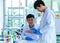 Asian young male scientist in white lab coat safety goggles and rubber gloves standing smiling showing green solvent in glass