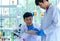 Asian young male scientist in white lab coat safety goggles and rubber gloves standing smiling showing green solvent in glass