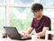 Asian young man wearing eye glasses sitting in the coffee shop looking  thoughtfully at screen of  computer laptop