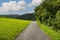 An asphalt road between a field of grass and trees, in the background hills overgrown with forest.