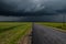 a asphalt road in a green field in front of a strong thunderstorm