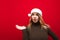Astonished attractive girl in warm sweater on christmas hat santa claus looks and points with her hand to the empty space,