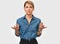 Astonished blonde woman gestures with both hands, wears blue denim shirt, posing against white studio background