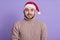 Astonished impressed young bearded man looking directly at camera with big eyes, wearing red santa claus hat and warm beige