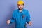 Astonished shocked young builder wearing yellow helmet and blue uniform, standing with opened mouth and spreading hands, making