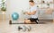 Athlete typing in smartphone, sitting on floor with glass of protein shake, with ball and dumbbells in interior of